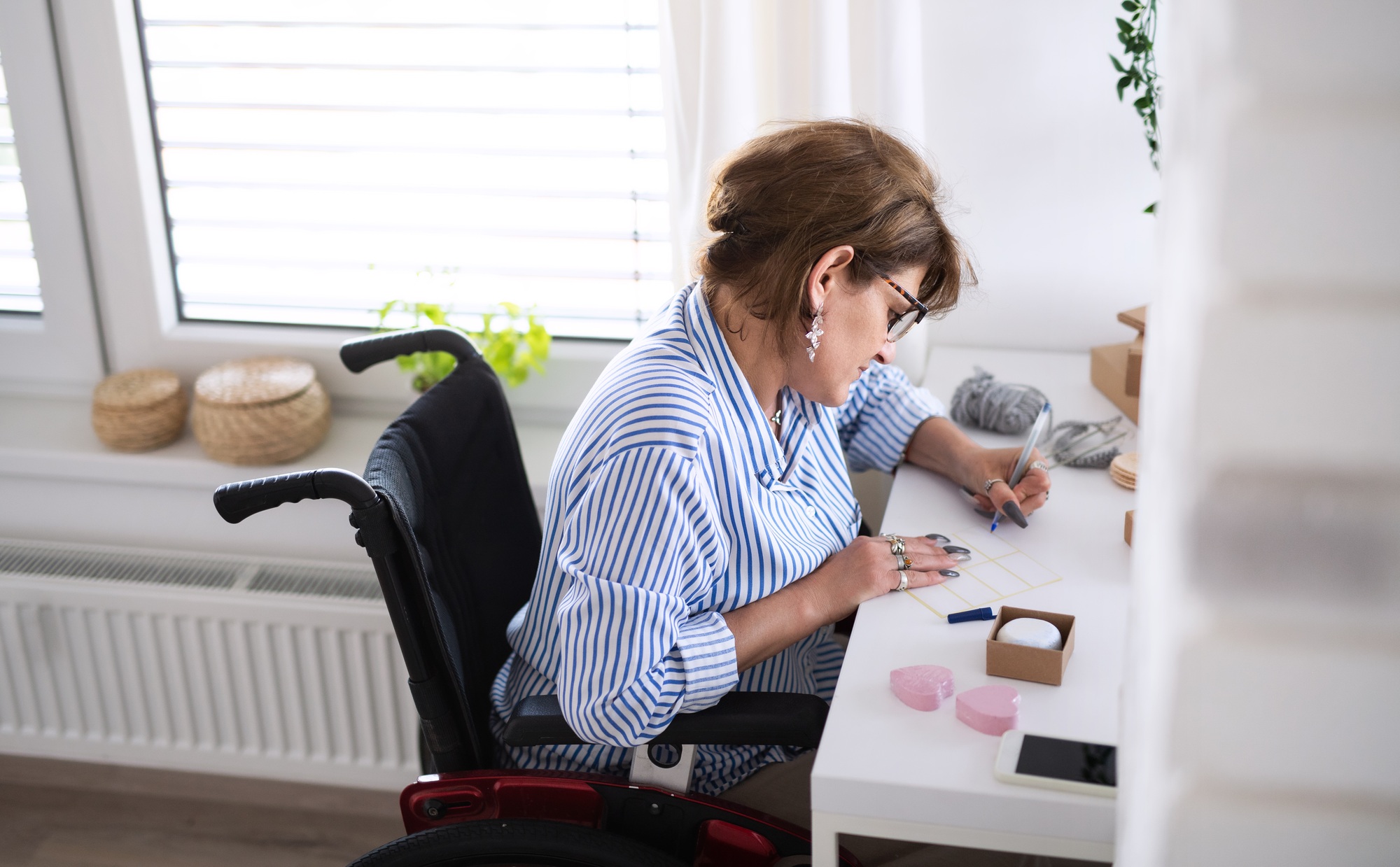Disabled mature woman in wheelchair packing handmade products indoors at home.