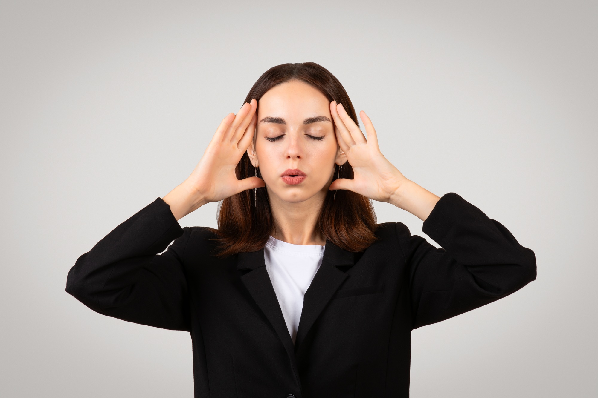 Focused businesswoman in a black blazer stands with eyes closed and hands on temples