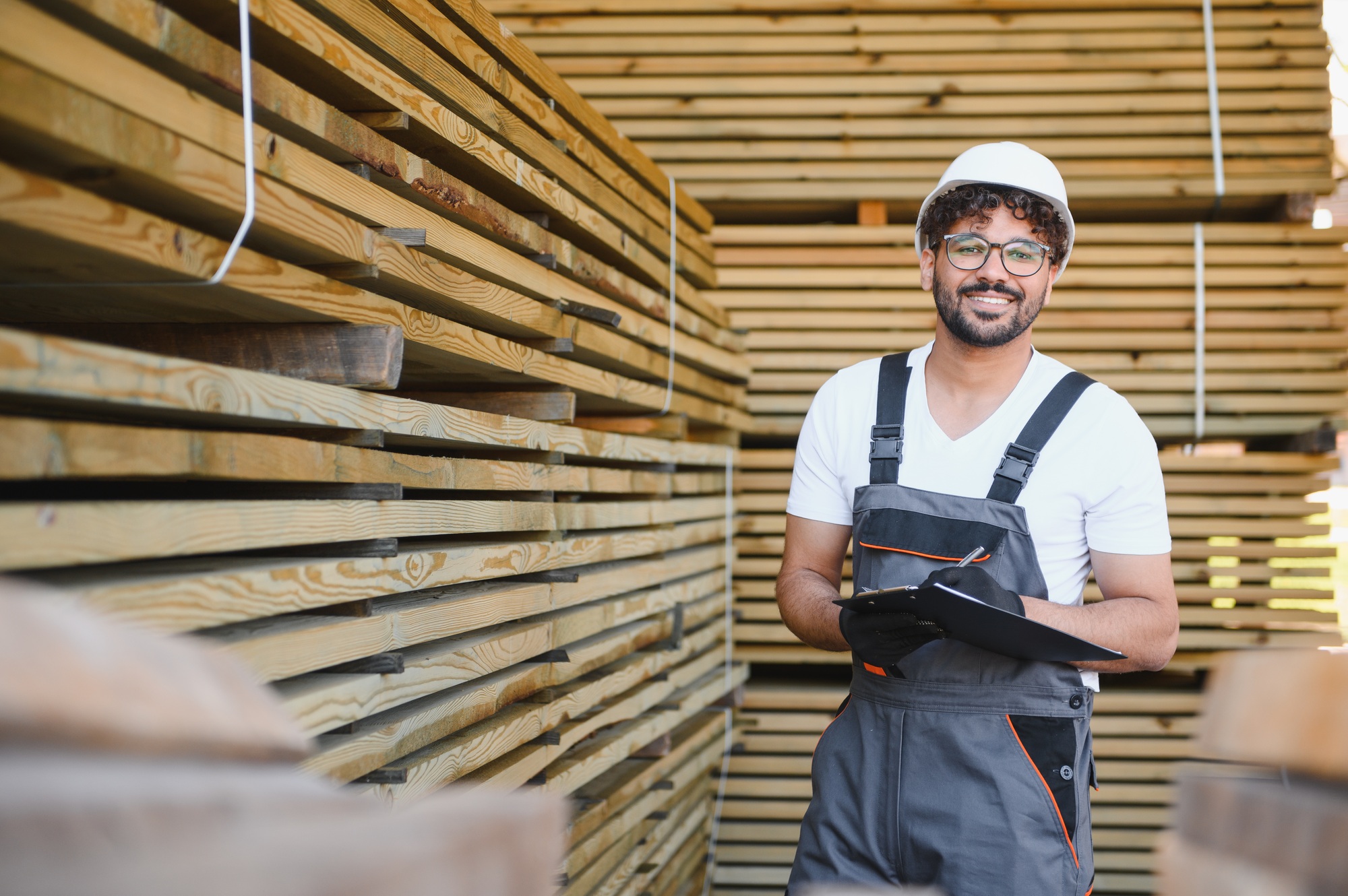 Smiling carpenter holding clipboard, checking lumber inventory in warehouse