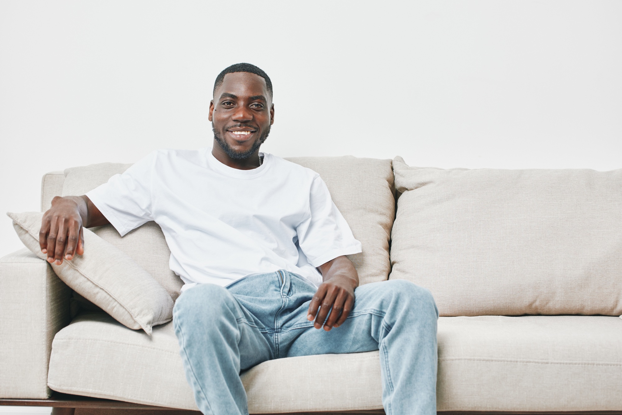 Smiling young Black man relaxing on a beige couch, wearing a casual white t shirt and light blue