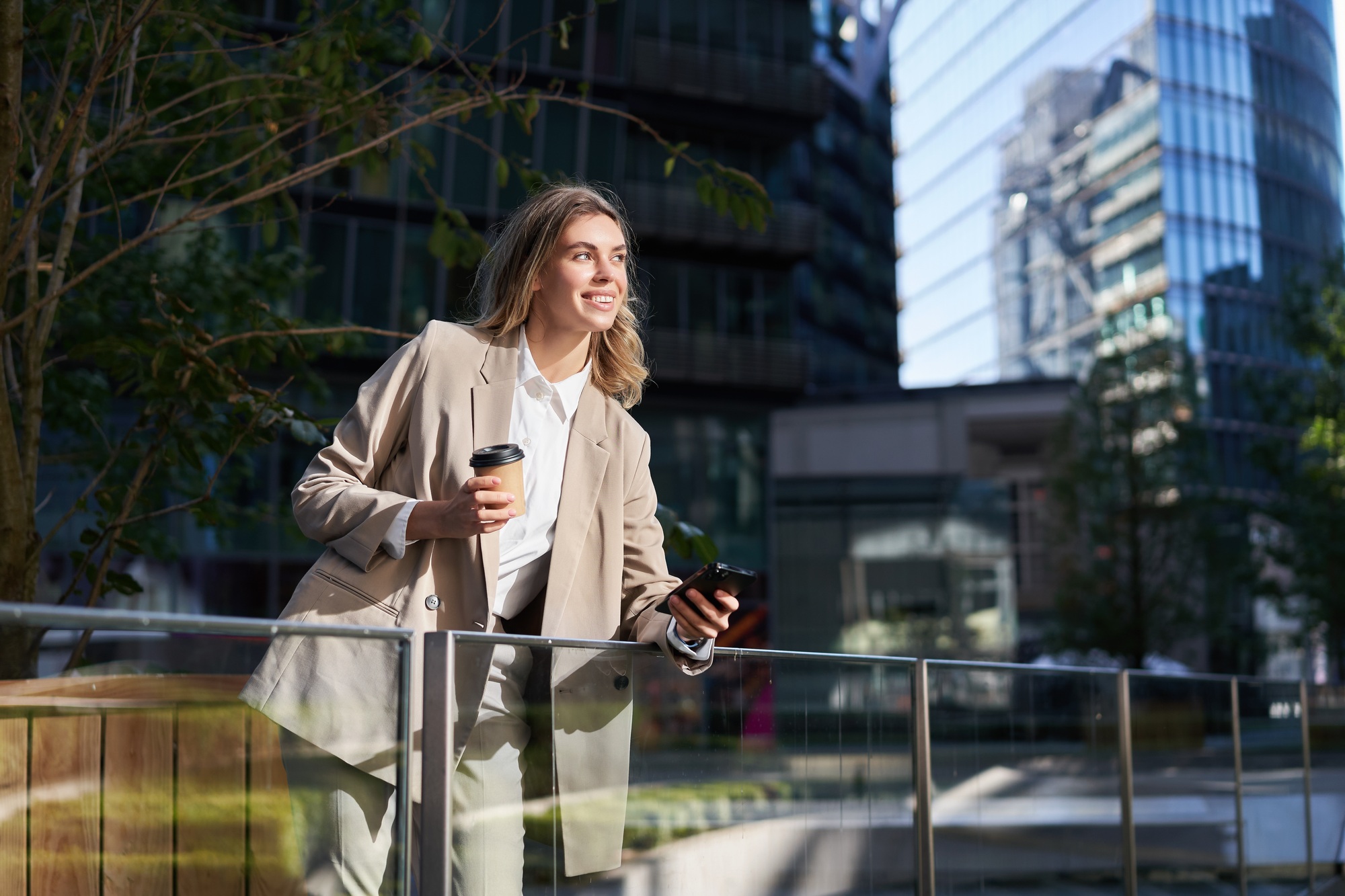 Successful businesswoman in suit, drinks her coffee and uses mobile phone, standing on street