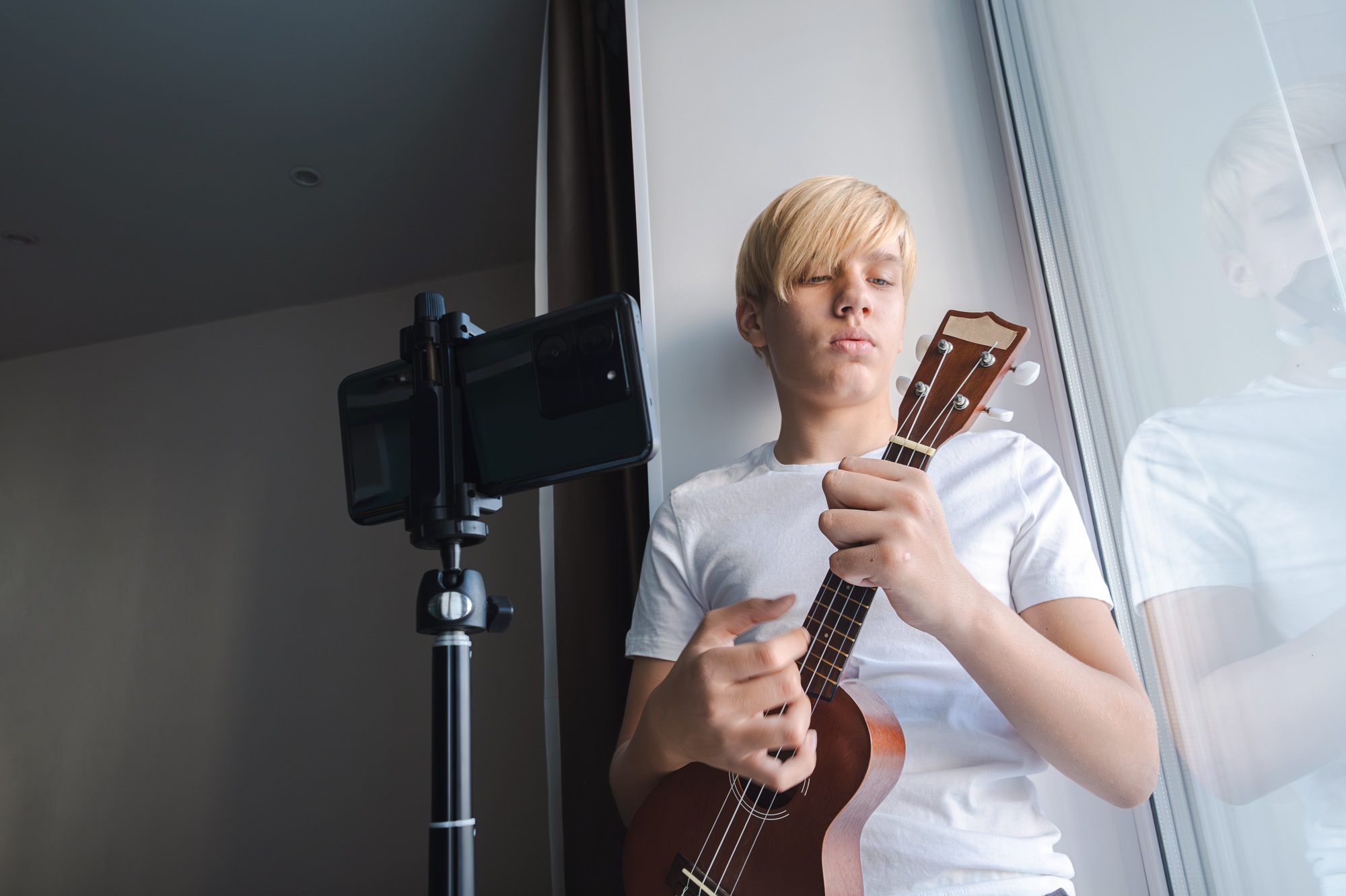 Teen playing ukulele while recording video on smartphone in natural lighting.