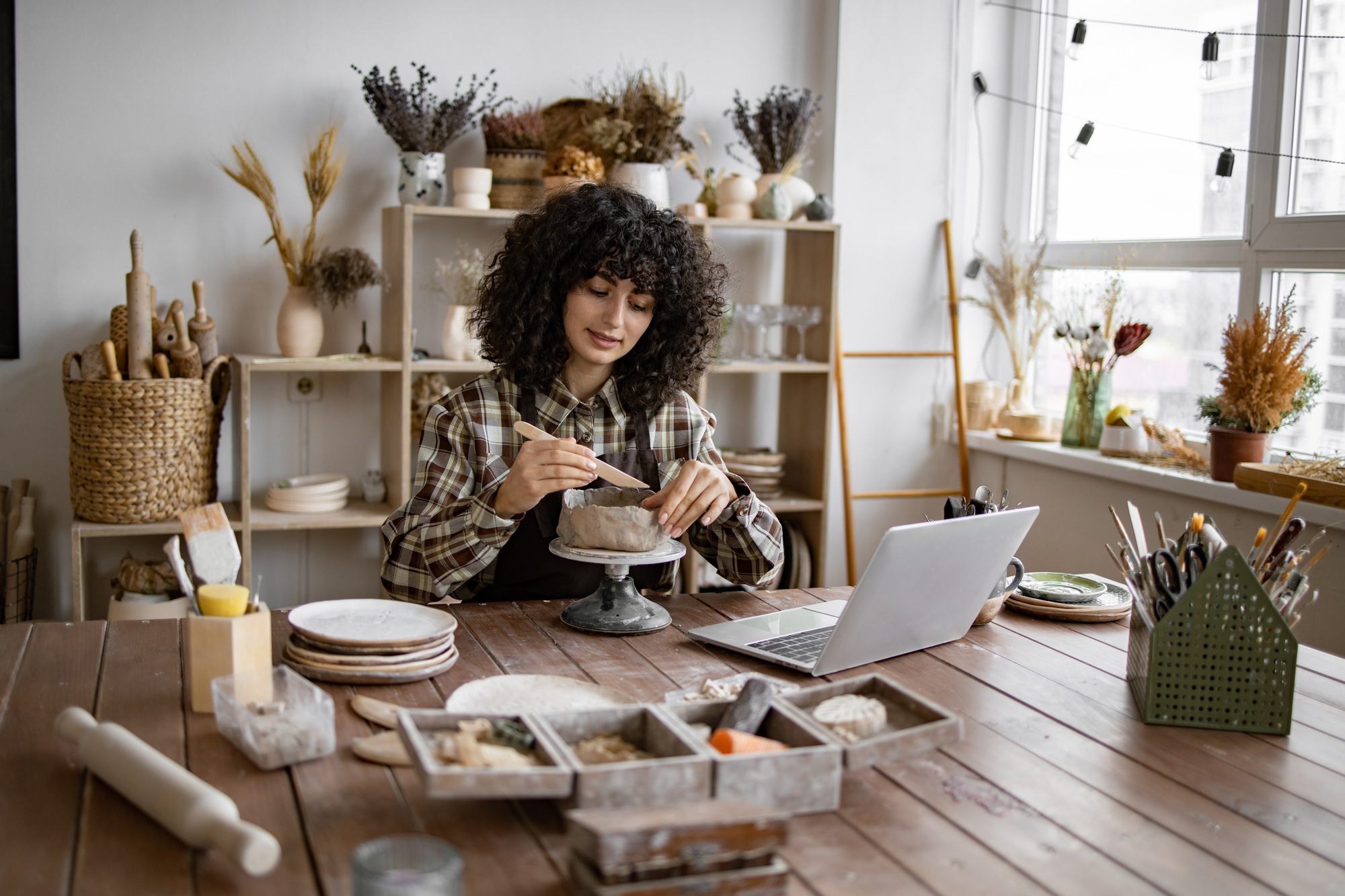 Woman crafting pottery in home studio with laptop