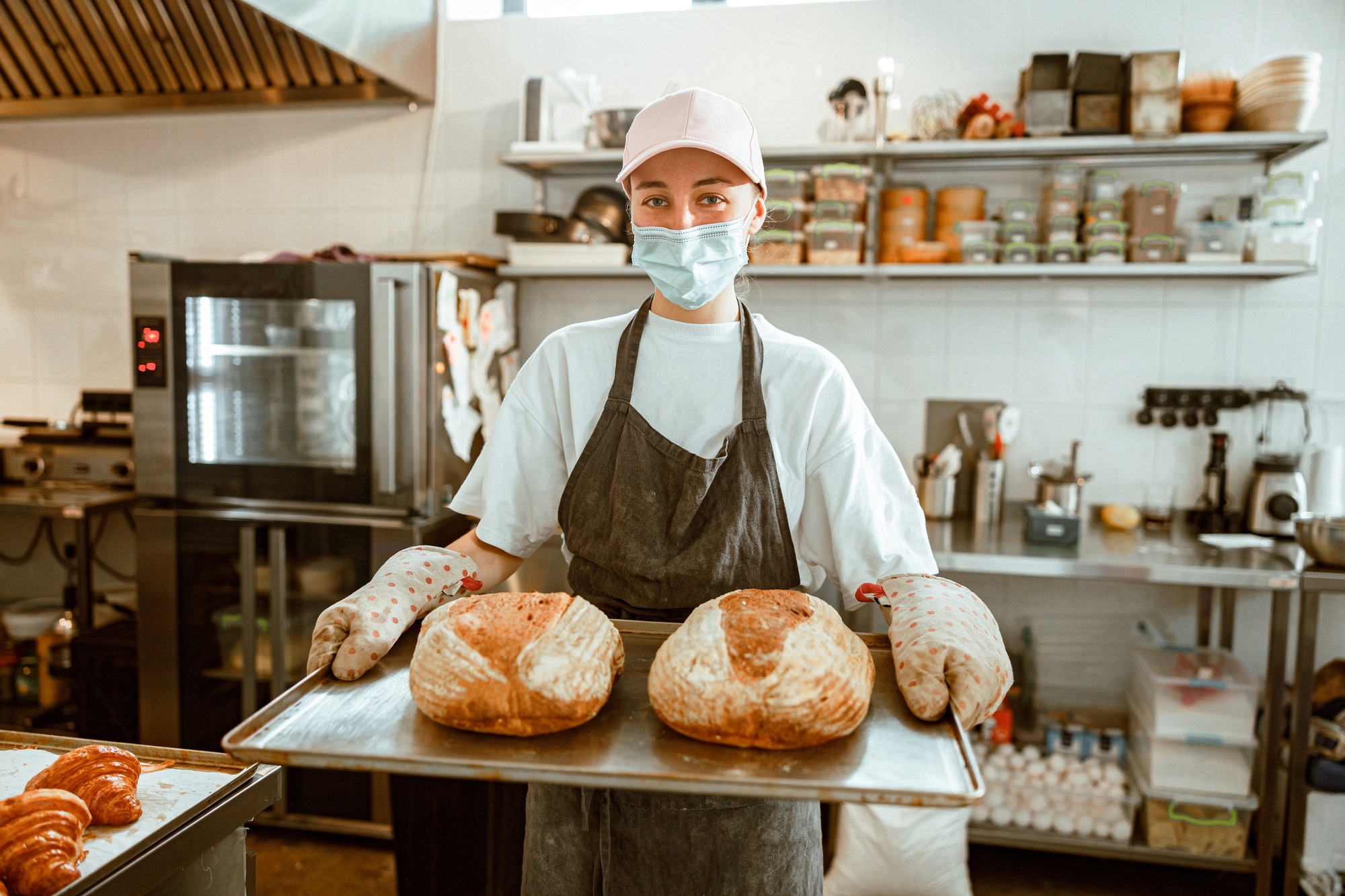Woman with medical mask holds tray with freshly baked breads in craft workshop