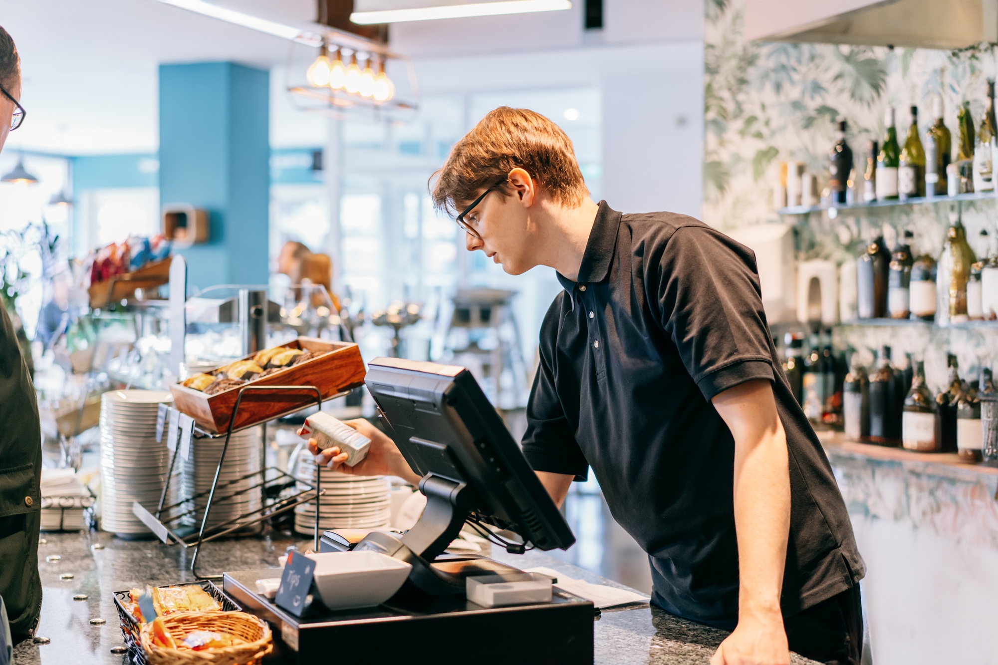 Young waiter serving customer at cash point in cafe. Man working with POS terminal. Cashier, barista