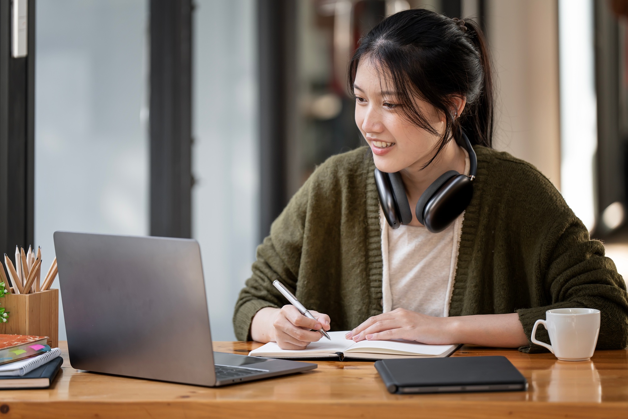 woman wearing headphones using laptop in cafe, writing notes, female student learning online