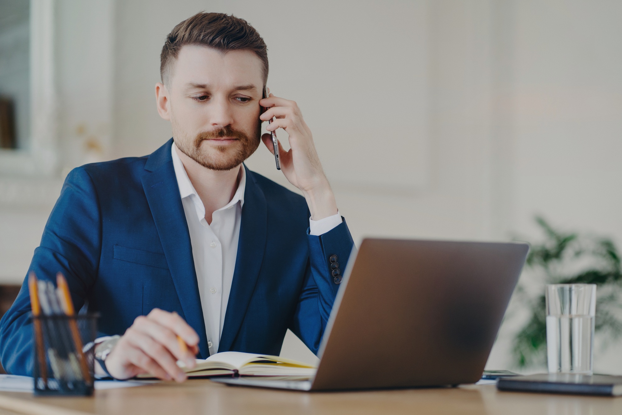 Serious businessman poses in office at desktop makes phone call concentrated at laptop